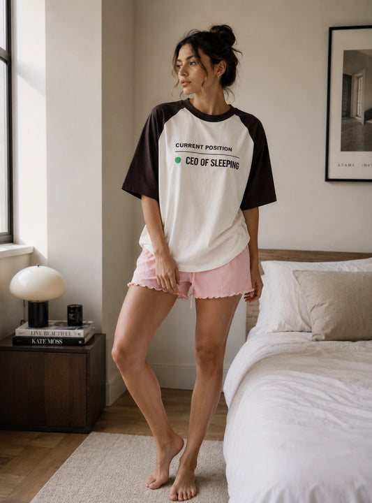 A woman stands barefoot in a bedroom wearing a raglan-style T-shirt with black sleeves and a white body, printed with the text 'CURRENT POSITION CEO OF SLEEPING' in black and green. She is also wearing light pink shorts with a scalloped edge.