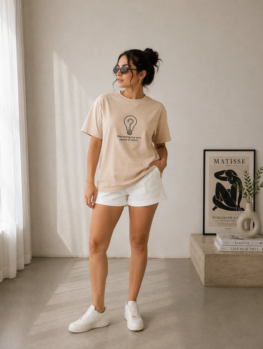 A woman modeling a beige graphic t-shirt with a lightbulb design, paired with white shorts and white sneakers, standing in a bright, modern room with a framed Matisse print on the wall.