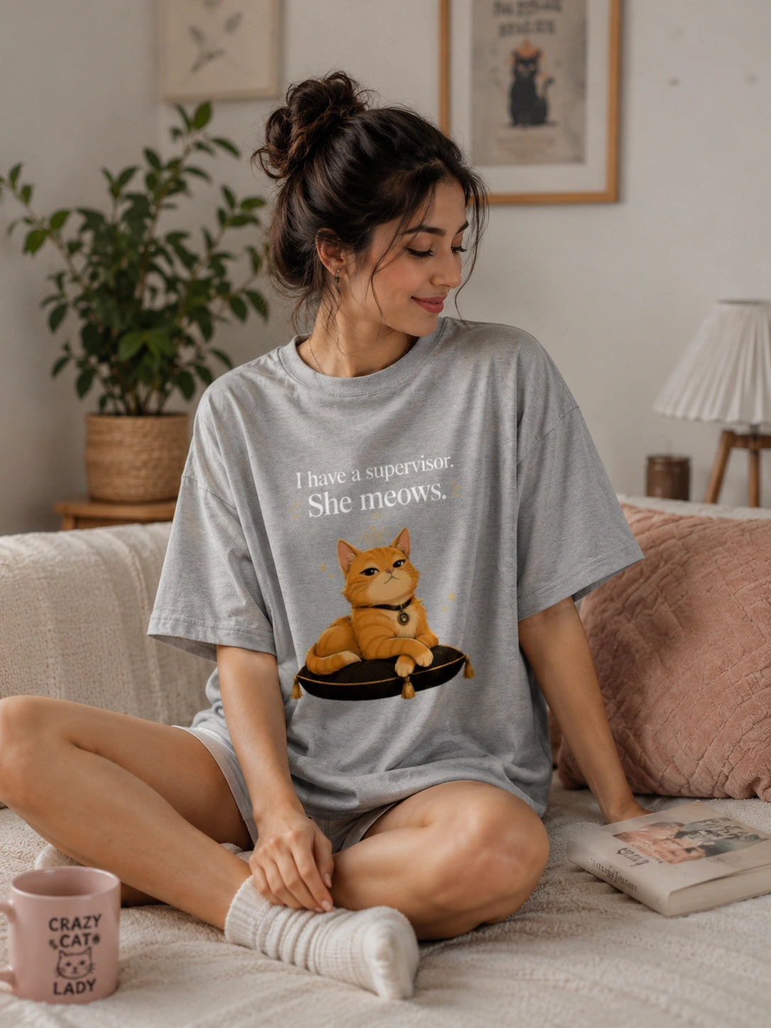 A woman sitting on a couch wearing a light gray graphic t-shirt with a cat illustration and the text 'I have a supervisor. She meows.