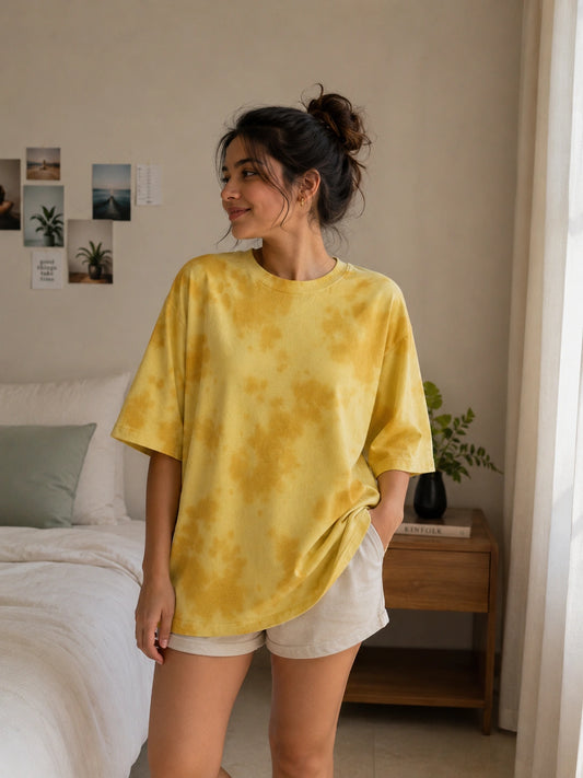 A woman wearing a yellow tie-dye oversized t-shirt and white shorts, standing in a bedroom setting.