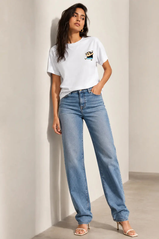 A woman modeling a white short-sleeve graphic t-shirt and light-wash blue straight-leg jeans, standing in a minimalist beige room corner.