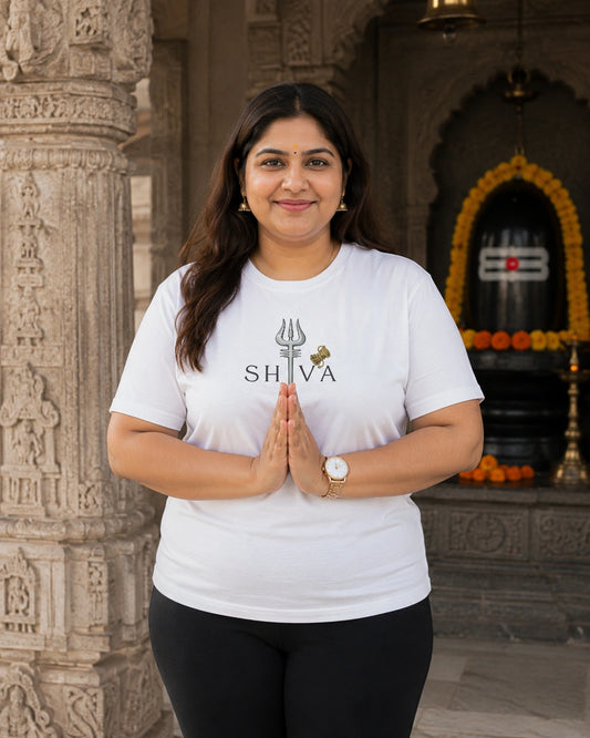 A woman wearing a white graphic t-shirt with a black 'SHIVA' trident logo stands in front of a temple backdrop with her hands pressed together in a prayer pose.
