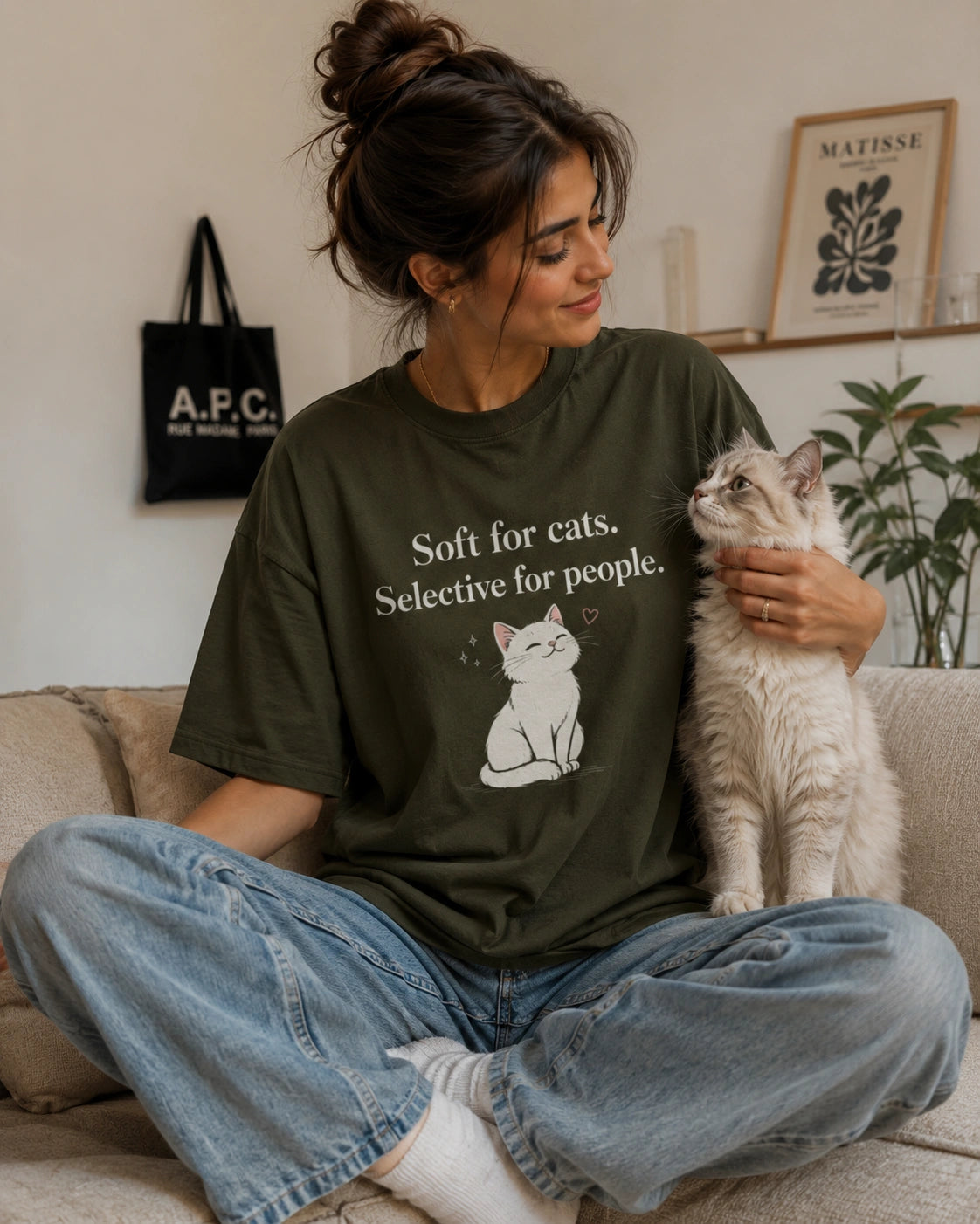 Woman wearing a olive green t-shirt with a cat graphic and text, sitting on the floor with a cat in a cozy living room.