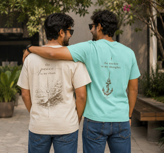 Two men standing outdoors with their backs to the camera, wearing matching graphic t-shirts. The man on the left wears a light beige t-shirt with a dark brown compass design and the text 'the peace in my chaos'.