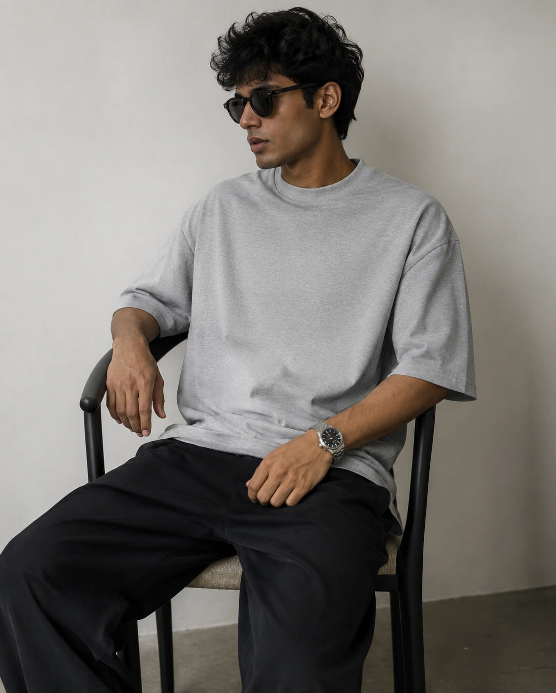 A man wearing a light gray oversized crewneck t-shirt and black pants, sitting on a black chair against a neutral off-white wall.