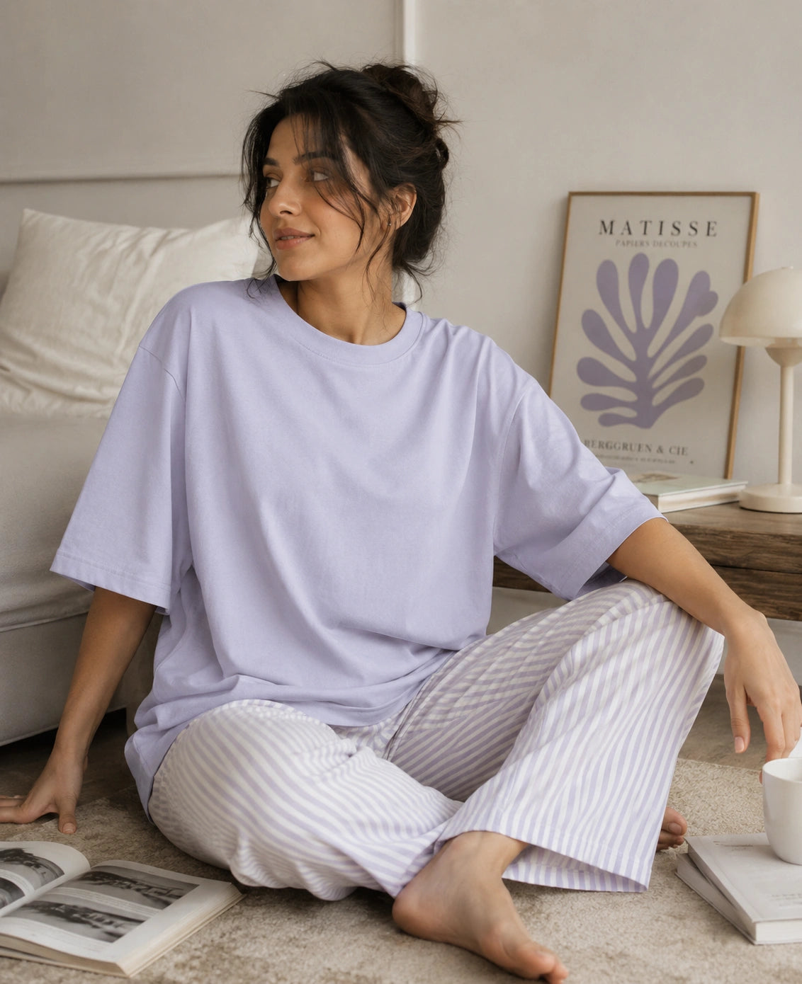 A woman sits barefoot on a beige carpet in a bedroom setting, wearing an oversized light lavender t-shirt and matching light lavender and white vertically striped pajama pants.