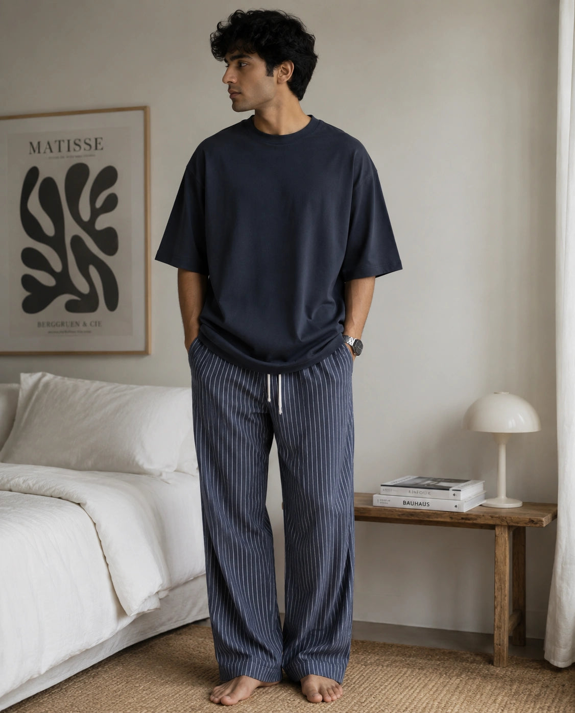 A man stands barefoot in a bedroom wearing a navy blue short-sleeve t-shirt and matching navy pinstriped lounge pants with a white drawstring. The room features a white bed, a wooden side table with a white lamp, and a framed Matisse print on the wall.