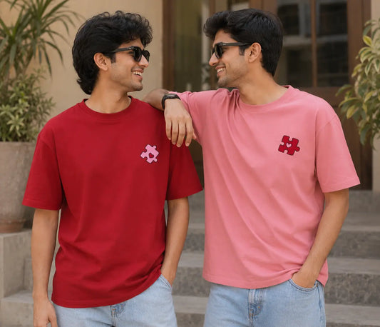 Two men wearing matching short-sleeve t-shirts with a small pink puzzle-piece logo on the left chest, standing on outdoor stone steps.