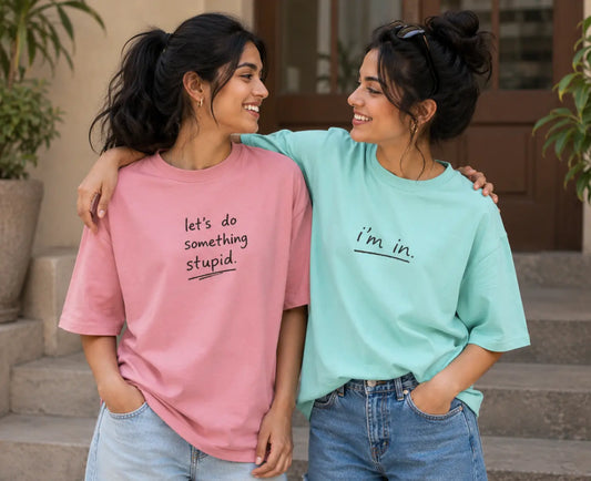 Two women standing on outdoor stone steps wearing oversized graphic t-shirts. The woman on the left wears a pink t-shirt with the black text 'let's do something stupid', and the woman on the right wears a mint green t-shirt with the black text 'i'm in.