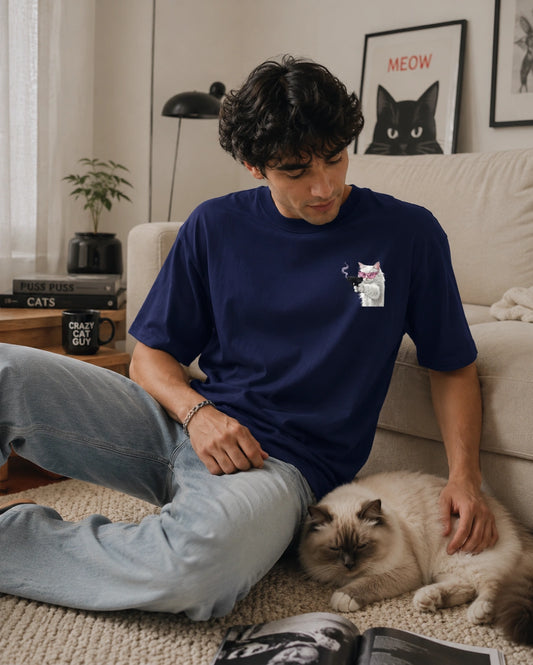 A man sitting on a beige carpet in a living room, wearing a navy blue t-shirt with a small cat graphic on the left chest and light blue jeans, petting a fluffy cream-colored cat.