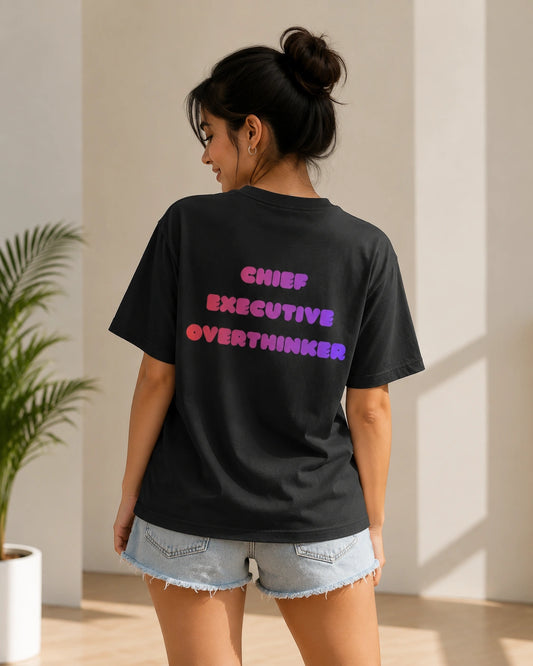 A woman shown from behind wearing a black graphic t-shirt with the text 'CHIEF EXECUTIVE OVERTHINKER' in a pink-to-purple gradient, paired with denim shorts in a brightly lit indoor setting.