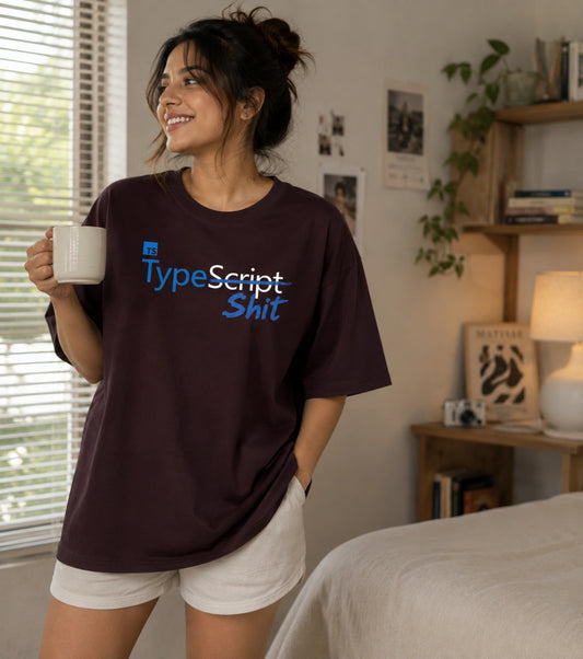 A woman stands in a bedroom holding a white mug, wearing an oversized dark plum T-shirt with the text 'TypeScript Shit' printed across the chest in blue and white, paired with white shorts.