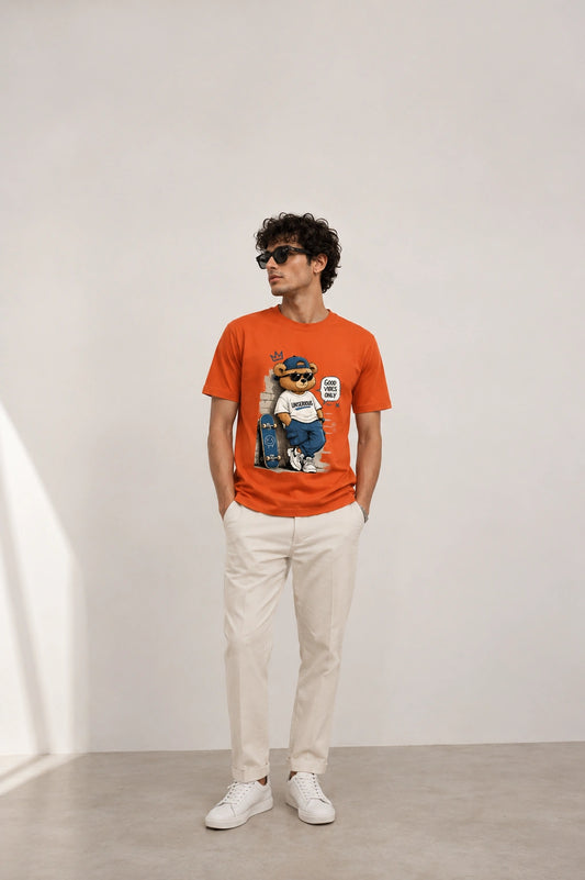 A man wearing a bright orange graphic t-shirt with a cartoon dog print, paired with off-white trousers and white sneakers, standing in a minimalist studio setting.