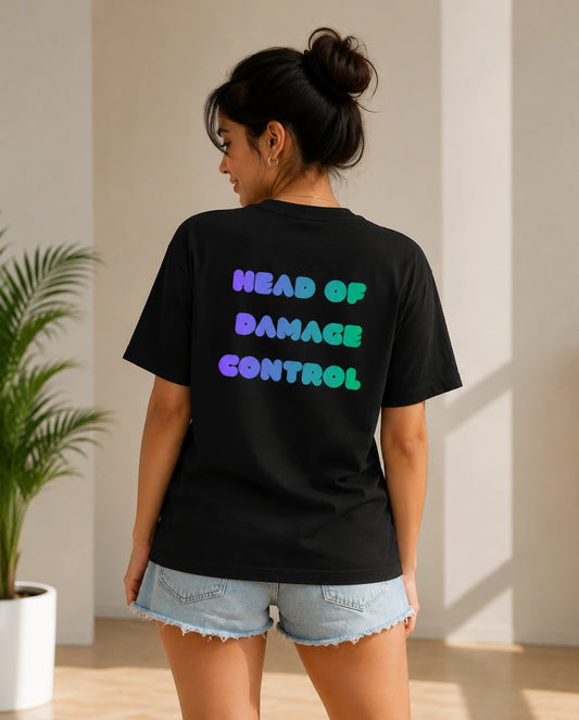 A woman shown from the back wearing a black graphic t-shirt with the text 'HEAD OF DAMAGE CONTROL' in a gradient of purple, blue, and green. She is wearing light blue denim shorts in a brightly lit indoor setting with a potted plant on the left.