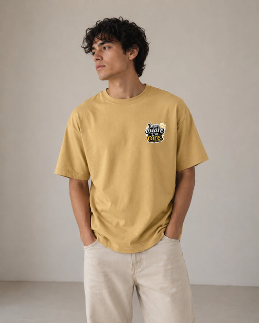 A man wearing a mustard yellow t-shirt with a 'Tiger Balm' logo on the left chest, paired with light beige pants, standing against a neutral beige wall.