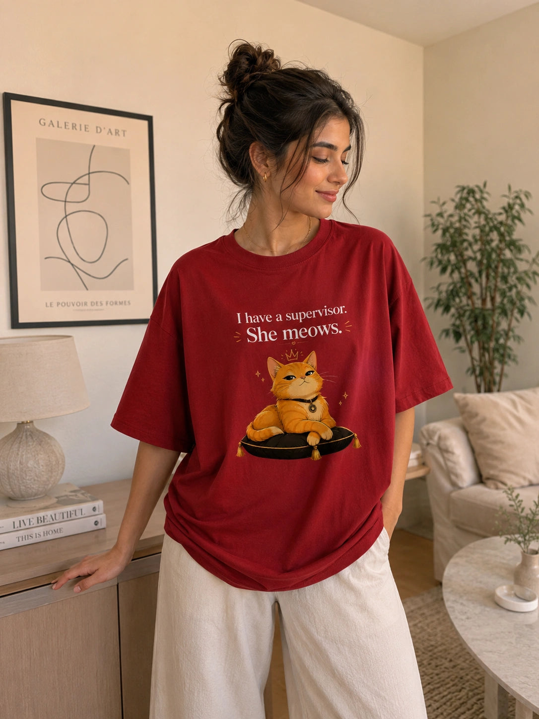 A woman wearing a red graphic t-shirt with a cartoon cat and the text 'I have a supervisor She meows.' in a cozy, modern living room setting.