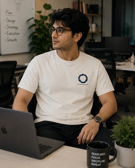 A man wearing a white crew-neck t-shirt with a small blue circular logo on the left chest sits at a desk in an office environment, looking at a laptop.
