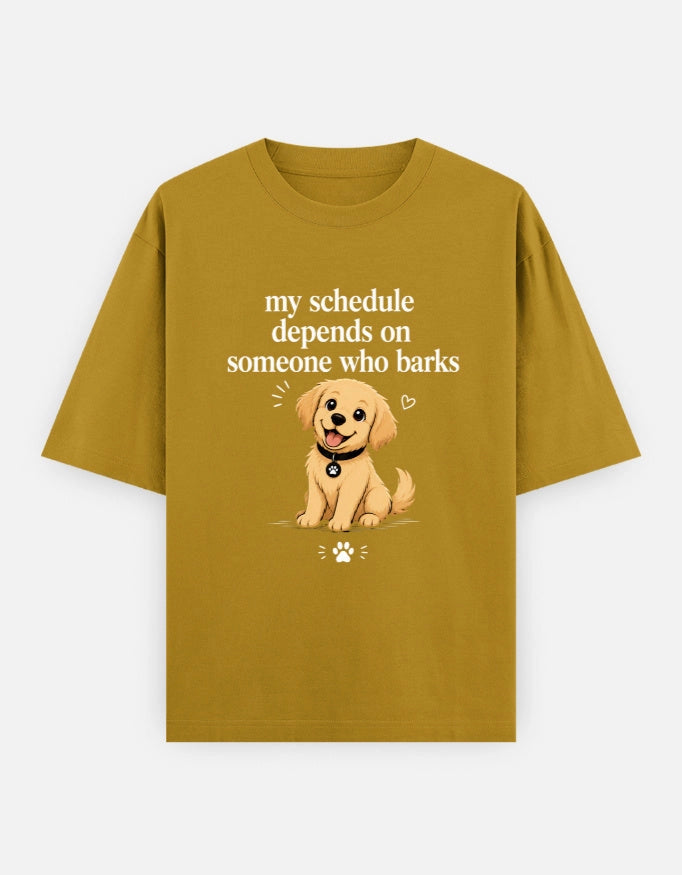 Unisex oversized T-shirt by The Unserious Studio featuring humorous "My Schedule Depends on Someone Who Barks" graphic on front.