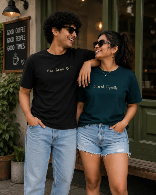 A man and a woman standing together on a street in front of a cafe, wearing matching graphic t-shirts. The man wears a black t-shirt with the text 'One Brain Cell' in white, and the woman wears a dark teal t-shirt with the text 'Shared Equally' in white.