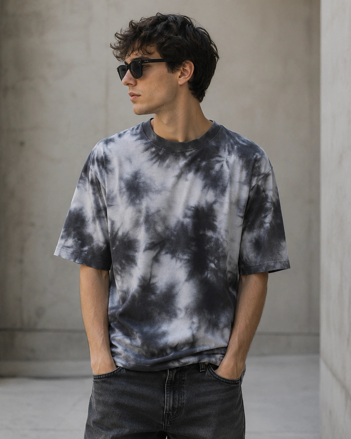 A man wearing a black and white tie-dye t-shirt and dark sunglasses, standing with his hands in his pockets against a concrete wall background.