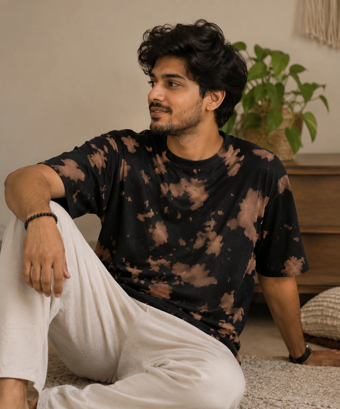 A man sitting on a carpet in a cozy indoor setting, wearing a black short-sleeve t-shirt with a brown tie-dye pattern and loose white pants.