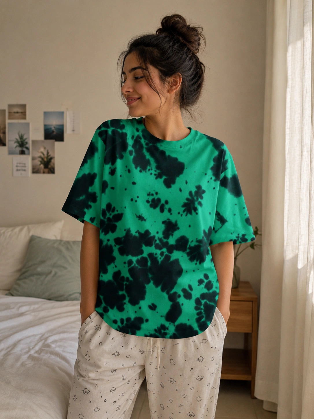 A woman wearing a green and black tie-dye t-shirt and white patterned pajama pants, standing in a bedroom setting.