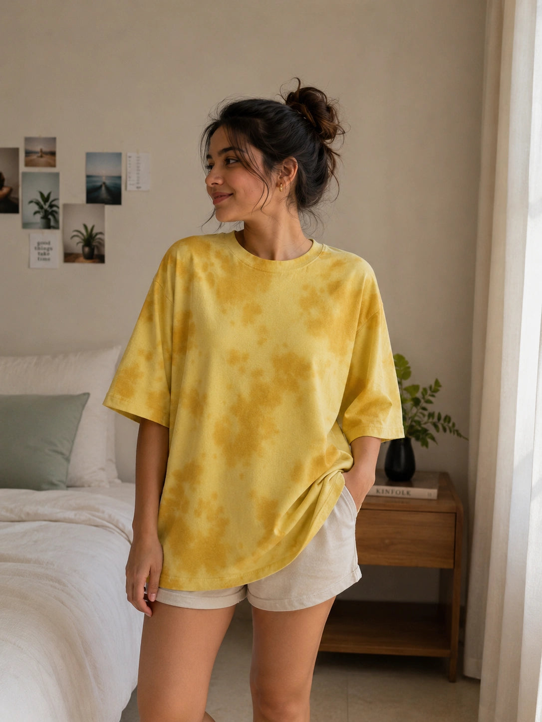 A woman wearing a yellow tie-dye oversized t-shirt and white shorts, standing in a bedroom setting.