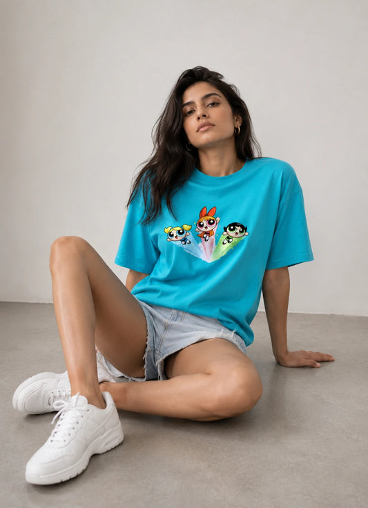 A woman sitting on a concrete floor against a neutral wall, wearing a bright turquoise blue t-shirt with a colorful graphic of the Powerpuff Girls on the front, paired with denim shorts and white sneakers.