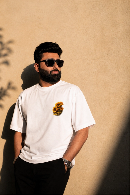 Man wearing a white t-shirt with a logo, sunglasses, and black pants against a beige wall.
