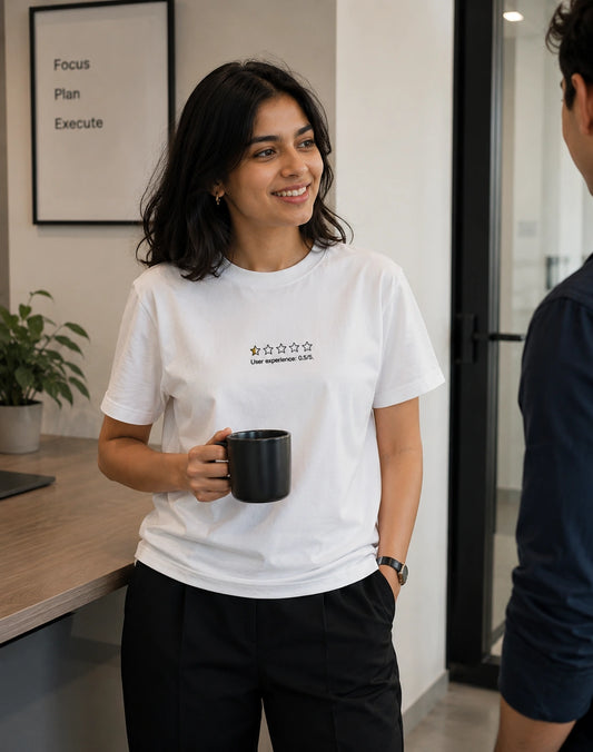 A woman in a white crewneck t-shirt with a black graphic logo on the chest, holding a black coffee mug in a modern office setting.