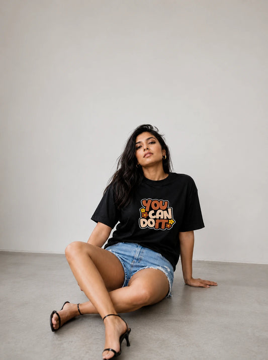 A woman sits on a concrete floor against a light gray wall, wearing a black graphic t-shirt with the text 'YOU CAN DO!' in orange, yellow, and white, paired with denim shorts and black strappy heels.