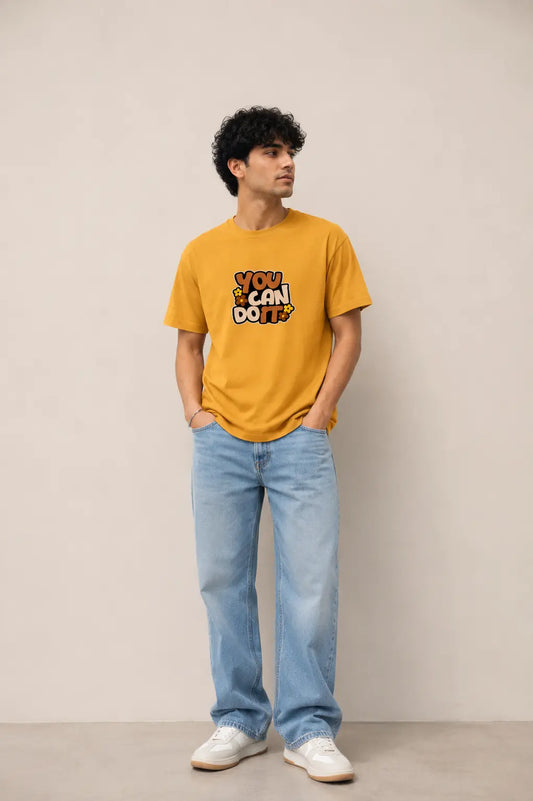 A man wearing a mustard yellow graphic t-shirt with the text 'YOU CAN DO!' in a stylized font, paired with light blue jeans and white sneakers, standing against a plain beige studio background.